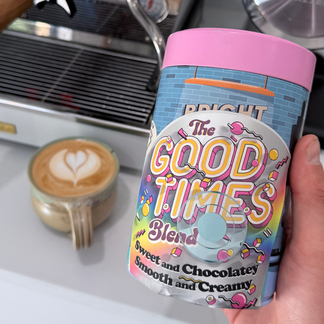 Canister labeled 'The Good Times Blend' held in front of a coffee cup with latte art.