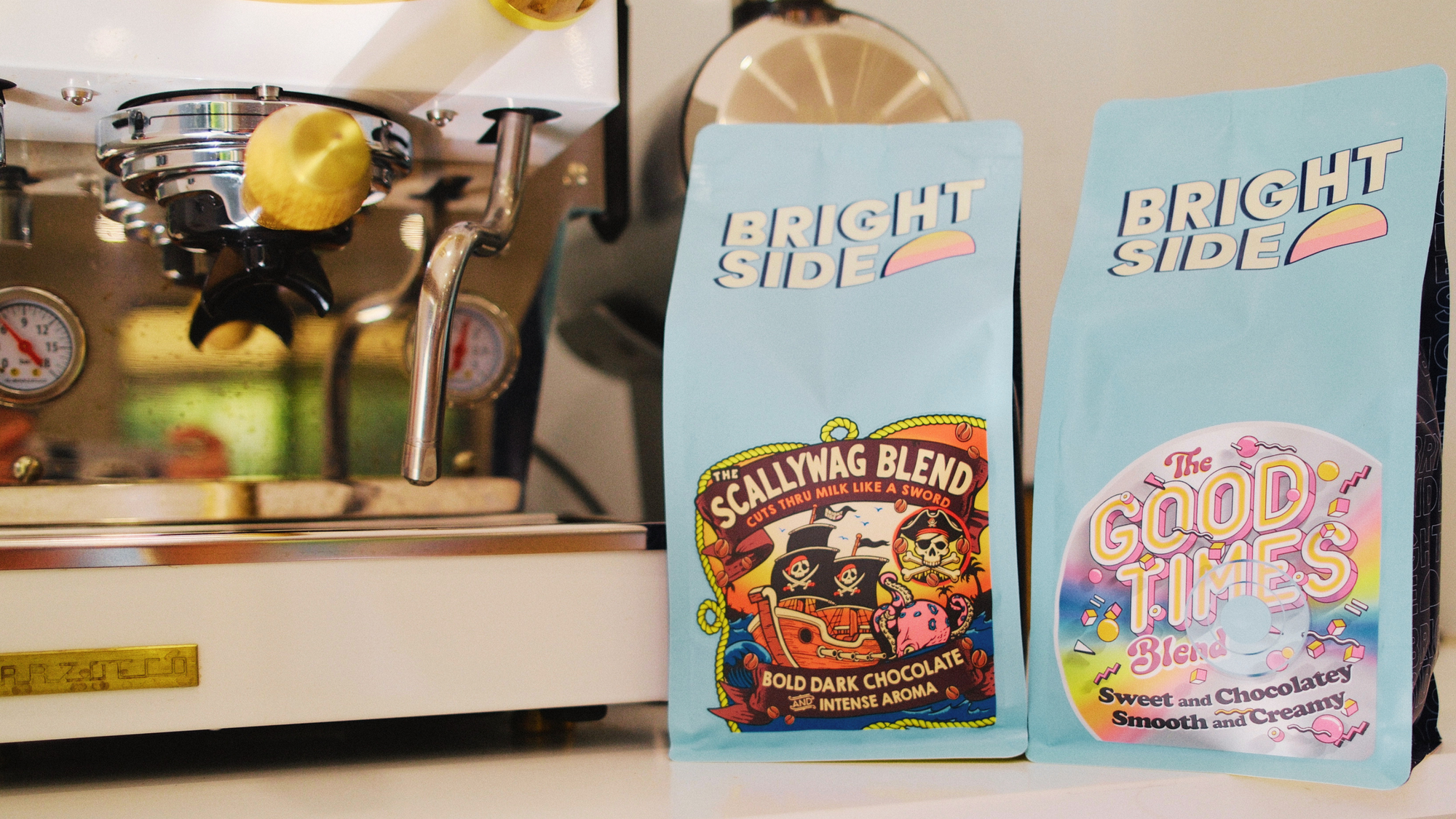 Two light blue bags of Bright Side coffee stands next to a stainless steel espresso machine on a countertop. The left bag is labeled 'Scallywag Blend' with a pirate illustration, and the right bag is labeled 'The Good Times Blend' with a colorful retro design.