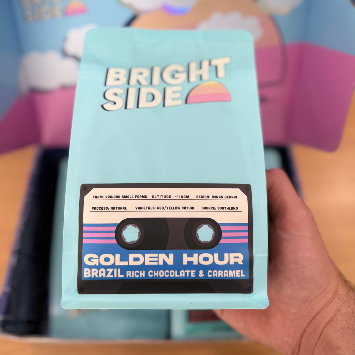 A hand holding a light blue bag of Bright Side Golden Hour Brazil Rich Chocolate & Caramel coffee in front of a colorful cardboard box.
