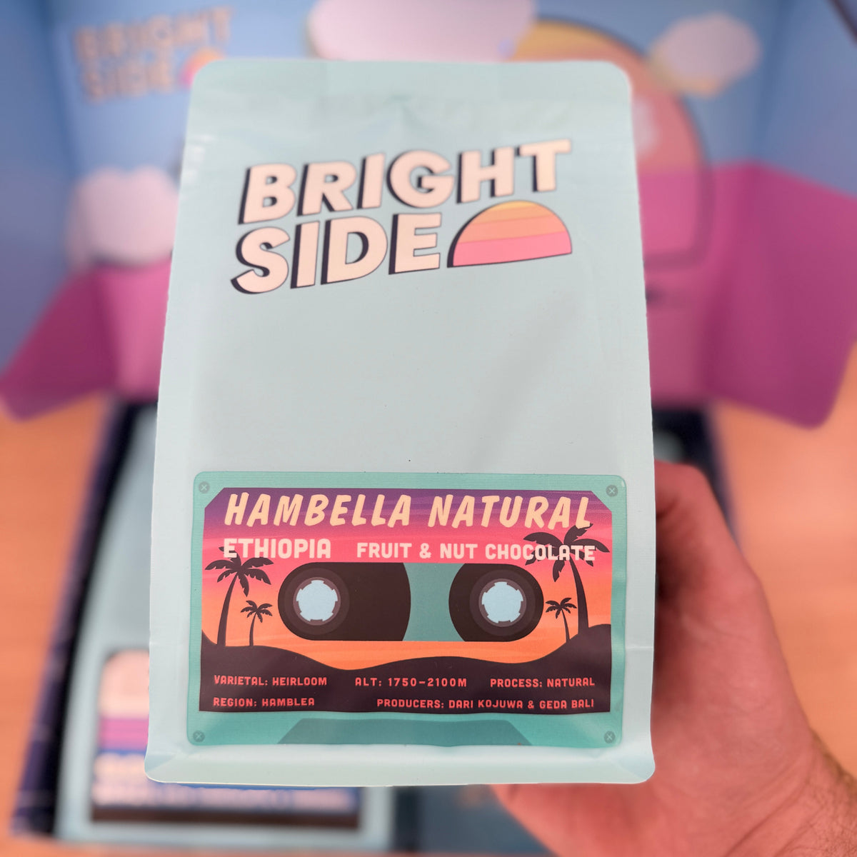 A hand holding a package of Bright Side Hambella Natural EthioPIR Fruit & Nut Chocolate against a colorful, blurred background.