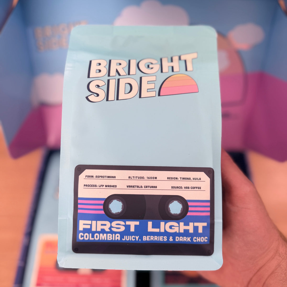 A hand holds a light blue coffee bag labeled 'BRIGHT SIDE' and 'FIRST LIGHT' in front of a matching branded box. The bag has a cassette tape graphic with colorful text and a small sunset illustration.